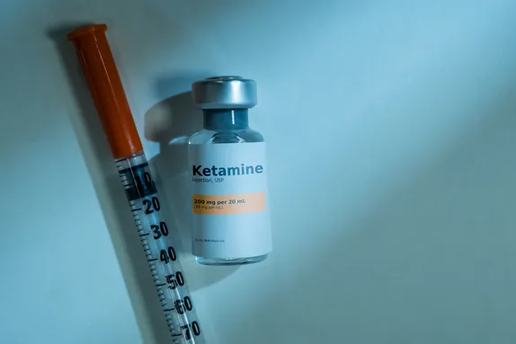 Mae ffiol wedi'i labelu “Ketamine” yn eistedd ar arwyneb glas golau wrth ymyl chwistrell gyda chaead oren.