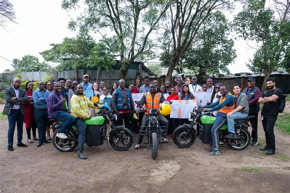 Group of people posing outdoors with three motorcycles, some holding posters and wearing safety vests.