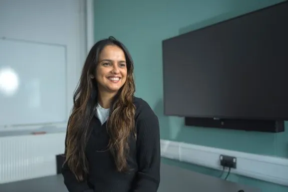 Chamathka is stood in a classroom in front of a large TV screen. She is wearing a black jumper and smiling for the camera.