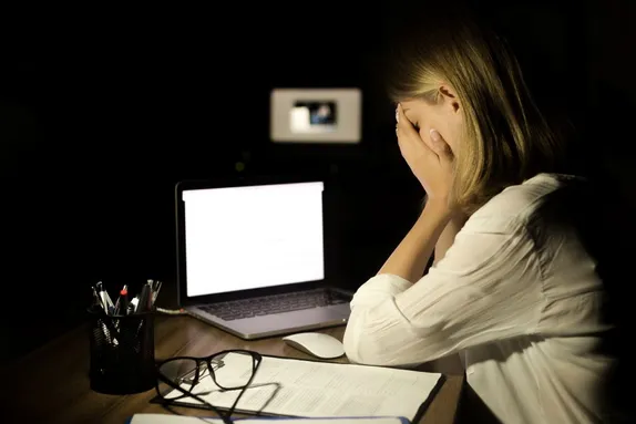 A person sits at a desk in a dark room at night, covering their face with both hands while looking toward a brightly lit laptop screen.