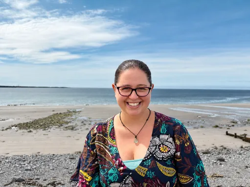 Heather smiles at the camera on a pebble beach with the sea and a blue sky behind her. The shoreline is stretching into the distance.