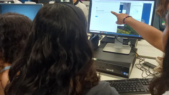 Three students looking at a computer screen, with one pointing to some coding shown on the screen.