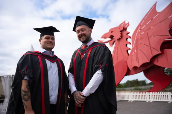 Ross Wills a Connor Jones, yn eu gwisg raddio a’u hetiau graddio, o flaen draig enfawr y tu allan i'r ICC yn Casnewydd, Cymru