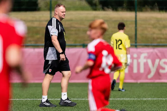A person in a black football kit walks across a football pitch in front of a goalpost. In the foreground, a player in a red kit is blurred in motion.