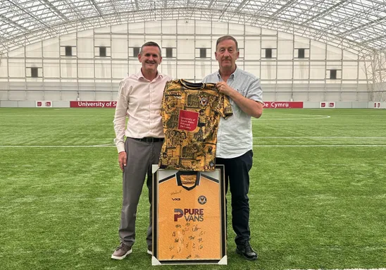 Two people stand on an indoor football pitch beneath a large arched training dome, holding a patterned Newport County football shirt featuring a University of South Wales logo. In front of them is a framed signed Newport County jersey.
