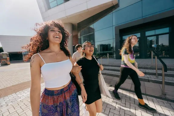 A group of students are walking outside a modern glass-fronted university building. Three people are prominent in the foreground carrying bags and backpacks as they move toward the entrance on a bright, sunny day.