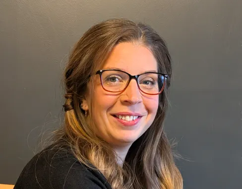 Jenny is facing the camera and smiling. She has long dark hair and wearing glasses.