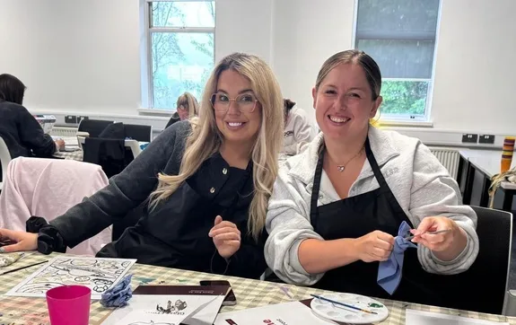 Two students sit together at a table during a wellbeing activity, there are paints and brushes on the table.