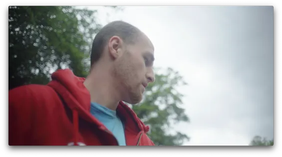 A man in a red hoodie is looking down at an angle with trees and sky visible behind him.