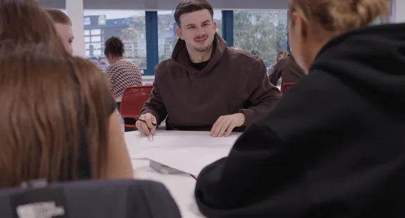 A group of built environment students wearing hoodies sit around a table discussing papers in the middle during a workshop at USW