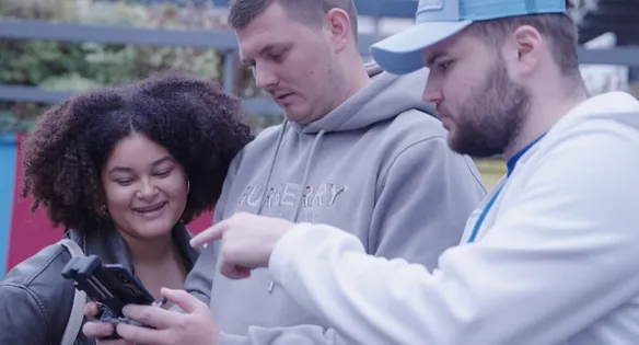 Three students in grey hoodies smile and chat while gathered around a piece of equipment at a building site