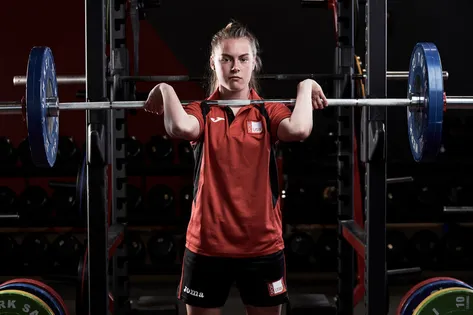 A student wearing USW Sports Kit is lifting a barbell with weights on either side.