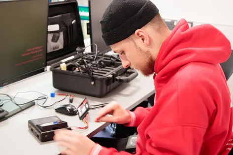 student in red hoodie with black hat, working at desk, handling electronic equipment