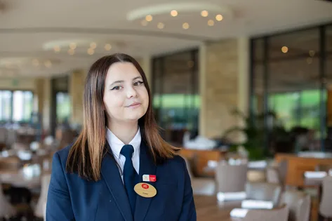 A student in hotel uniform is looking into the camera, they are in a dining area of Celtic Manor Resort