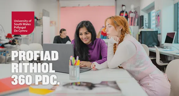 Two students are sat at a table in a fashion studio looking at a laptop together.