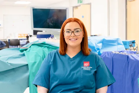 ODP student Hollie smiles directly at the camera whilst wearing her blue scrubs. There is medical equipment behind.