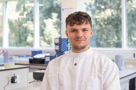 Biological Science student, Oliver, smiles for a profile shot in a science lab, wearing a white lab coat
