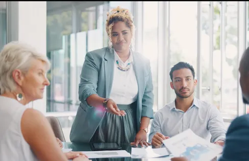 A group of business professionals sitting around a table. One woman is stood up, presenting ideas to the group.
