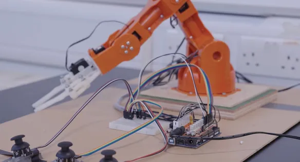 An orange robotic arm with white claws sits on top of a wooden desk with a motherboard, small plugs and wires in an engineering workshop at USW's Treforest campus