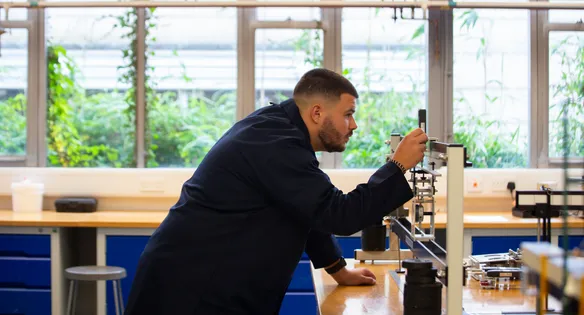 A student wearing navy engineering overalls adjusts the measurements on a piece of engineering equipment on top of a desk in a workshop at USW's Treforest campus. Behind the student is windows showing plants outside against a white wall