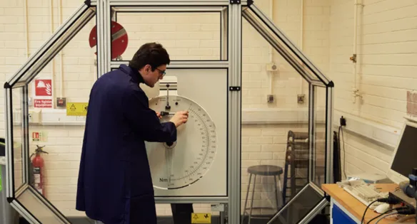 A mechanical engineering student wearing navy overalls with their back to the camera adjusts a piece of engineering equipment on the wall of an engineering workshop