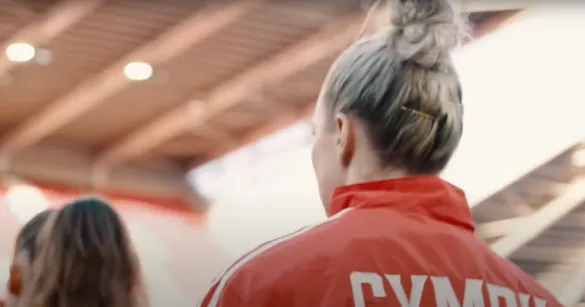 The back of a female footballer wearing a red jersey that says 'Cymru'. She has blonde hair tied in a bun