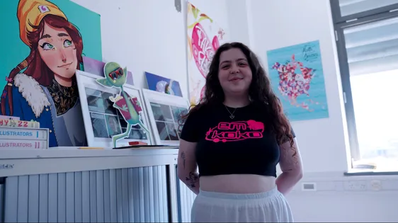 Illustration student smiles at the camera while stood with their hands behind their back in their bedroom with books and illustrations they've created behind them