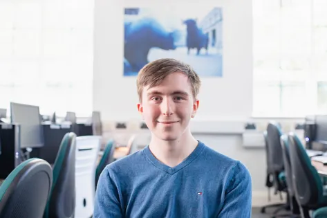 Studet Leo Humphreys smiles at the camera while wearing a blue t-shirt and sat in a computer room with various desktops on desks behind him