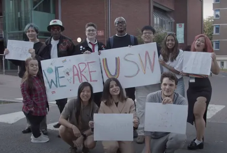 A group of students of different ethnicities and backgrounds smile while standing in a group outside Treforest campus and holding a handmade paper banner with colourful hand drawn writing saying 
