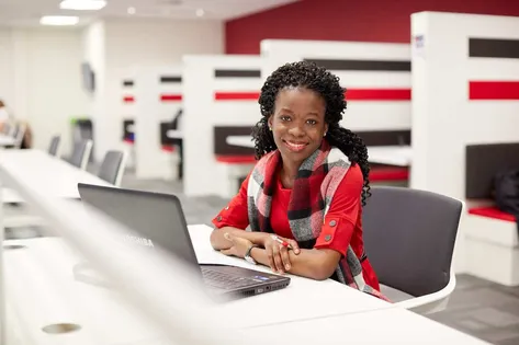 A student sat in the library working on a laptop and smiling at the camera.