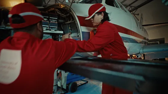 Two students in red uniforms collaborating on an aeroplane, focused on their tasks with tools in hand.