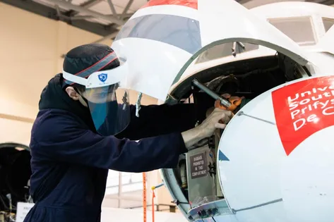 A student wearing navy USW engineering overalls, a face mask and a face shield works on the front of the small plane which has its nose panel open inside the USW aerospace workshop at the Treforest campus