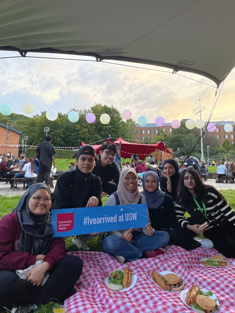 A group of international students sat on the grass in treforest campus with a welcome sign