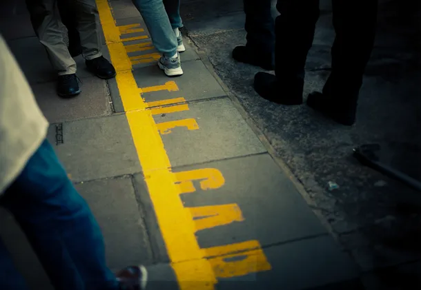 A close up of peoples feet walking along a busy train platform. The edge of the platform reads 