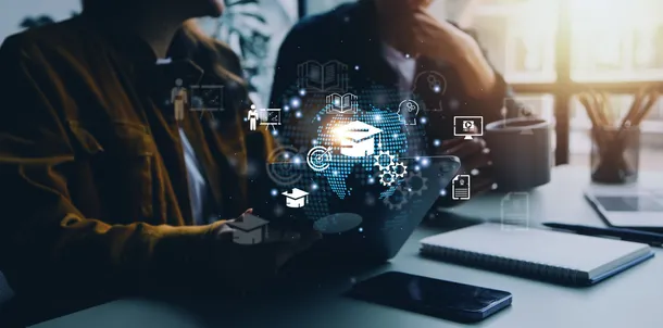 A shot from the head down of two people sat at a desk on a laptop. There are glowing icons in the foreground which symbolise leaning and technology.
