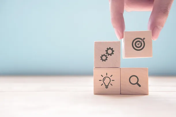 An image of four wooden building blocks with different icons on them; cogs, a dartboard, a magnifying glass and a lightbulb.