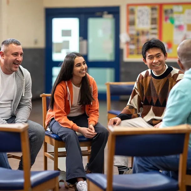 A group of people sit in a circle smiling and listening to each other talk in a community hall