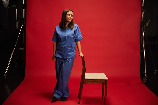 Nursing alumni, Megan, wearing blue scrubs stood in front of red backdrop holding on to the back of a chair
