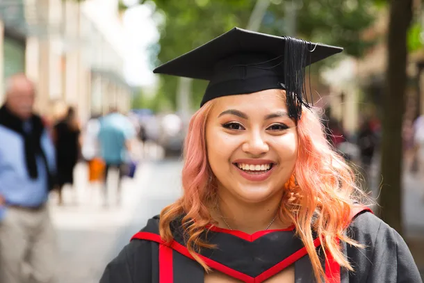 A close up of a student with long coral hair smiling while wearing black and red graduation robes and a graduation cap.