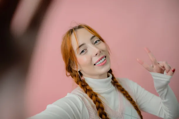 A girl is taking a selfie and making a peace sign against a pink wall.
