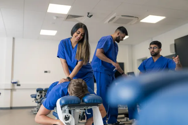 female student practising massage technique on male student, two male students in the background interacting