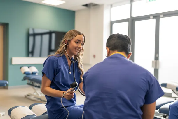 female student sat down with stethoscope, testing male student who has his back to the camera