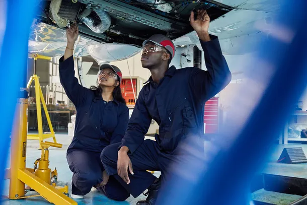 two students in navy overalls looking up at the underneath of a plane