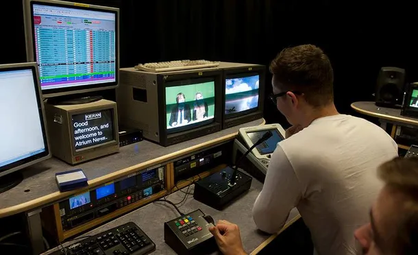 A student sat in the tv studio watching a tv screen and a teleprompter and talking to peers through the microphone.