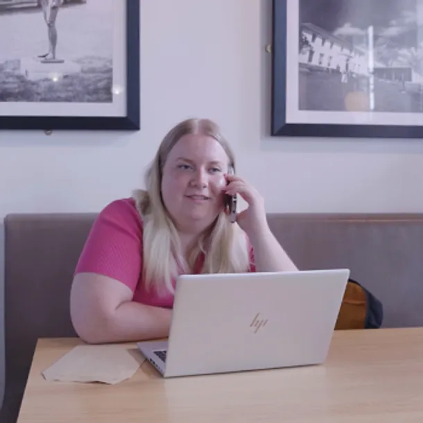 A blonde woman is sat at a table, talking on a mobile phone with a laptop in front of her.