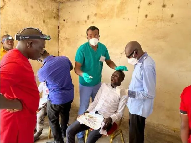 Albert Akhidenor performs dental surgery on a man in a chair surrounded by other dental staff in an old looking building with yellow walls