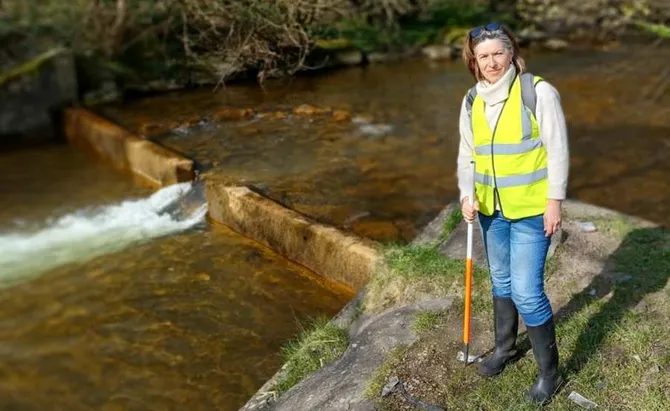 A woman in a high vis looking towards the camera standing next to a river