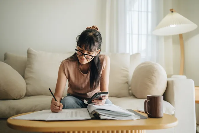 A young woman wearing glasses looking through a binder and using their phone to plan her monthly budget.