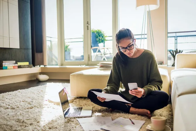 A person sat on the floor surrounded by papers and a laptop, using a calculator.
