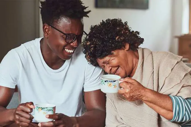 A mother and son are sat on a sofa drinking tea together and laughing.
